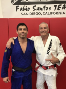 Two Brazilian Jiu-Jitsu practitioners, Justin Flores and Fabio Santos, one in a blue gi and the other in a white gi with a red and black belt, posing together in a dojo.